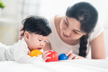 Obraz na płótnie Canvas Asian young mother look at baby, hold son feet and kiss with warm love while cute boy close his eyes playing peekaboo with mom on bed at home. Mother child relationship is good for kid development.