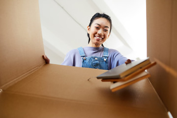 Low angle view at young Asian woman looking into cardboard box and smiling happily while packing or...