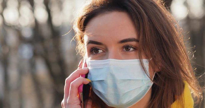 Portrait Woman dressed in orange red coat wearing Protective Face Mask talking mobile phone in the park . close up view. Sunny day Sun flayers pandemic disease virus. COVID-19 coronavirus infection