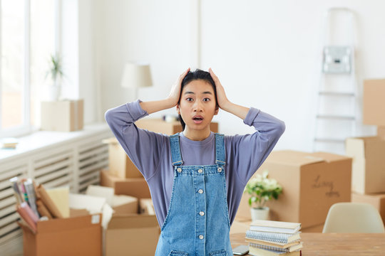 Waist Up Portrait Of Young Asian Woman Panicking While Standing Among Cardboard Boxes In Empty Room And Looking At Camera With Big Eyes, House Moving Or Relocation Concept, Copy Space