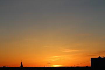 Sun below the horizon and the roof of the church with a cross, television antenna, roof of a multi-storey urban building in the background fiery dramatic orange sky at sunset or dawn backlit by the