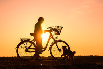 Obraz premium Senior woman standing near a bike and feeding a dog, silhouette of person at sunset with pet, rest at a trip 