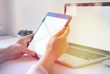 Close-up of tablet with blank screen in hands of young woman sitting at white table and touching screen. On table laptop with blank screen,cup of coffee and flowers. Minimalis. Mock up,space for ad
