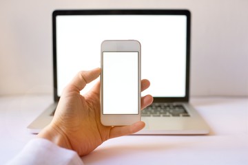 Close-up of smartphone with blank screen in hands of young woman sitting at white table and touching screen. On table laptop with blank screen,cup of coffee. White style. Minimal. Mock up,space for ad