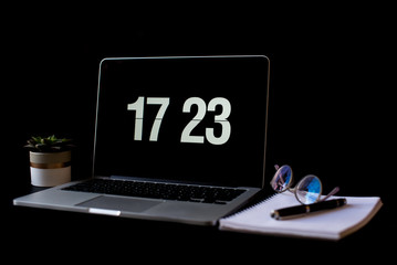 Close-up photo of laptop with time screensaver placed on black desk in room interior with glasses, notes, pen on the dark wall