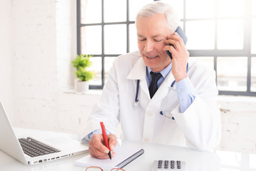 Senior male doctor using mobile phone and laptop while working in doctor's office