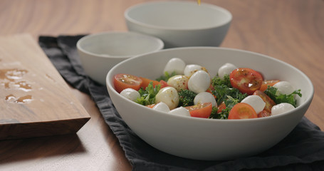 man hands pour dressing over salad with kale , cherry tomatoes and mozzarella in white bowl
