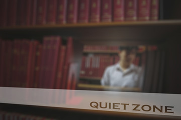 Opaque white frosted glass sticker with wording “QUIET ZONE” stick on clear mirror and blurry young man wearing white shirt selected books on bookshelf in a public library room background.