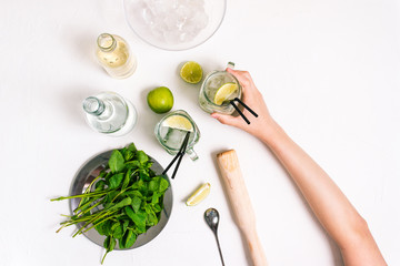 Girl holds a fresh mojito cocktail in her hand. Ingredients for making a summer drink on a white...