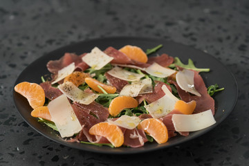 Salad with prosciutto, tangerines and vintage cheese on black plate
