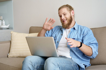 Happy young man waving hand video calling on laptop sitting on sofa working studying from home remote office. Smiling male distance worker student doing online conference webcam chat on computer.
