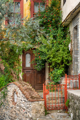 Entrance of a house with an ancient wooden door sunken in greenery by colored bushes and an olive tree in the old town of Kavala, northern Greece