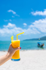 First-person view. Girl holds a glass cup of cold mango fresh on the background of a sandy tropical beach. White sand and a boat. Fairytale vacation in Thailand