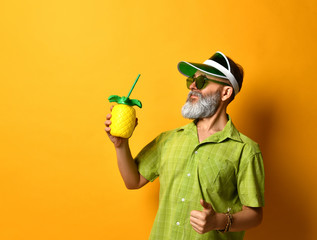 Gray-bearded grandpa in green sun visor, shirt, sunglasses. He smiling, holding cocktail bottle in form of pineapple with tube, clenched fist, posing on orange background.