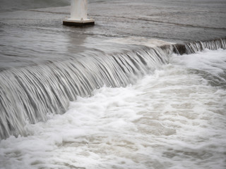 Close up view of water cascading or waterfalling over a concrete pier edge in Lake Michigan in Chicago during a windy day as waves crash into the shoreline.