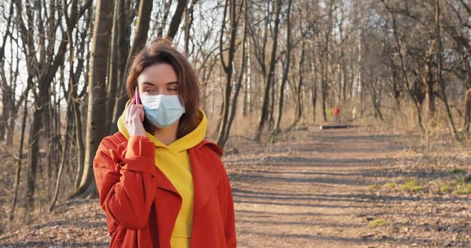 COVID-19 coronavirus infection Woman dressed in orange red coat wearing Protective Face Mask walking in the park with dog. Sunny day Sun flayers pandemic disease virus epidemic air.