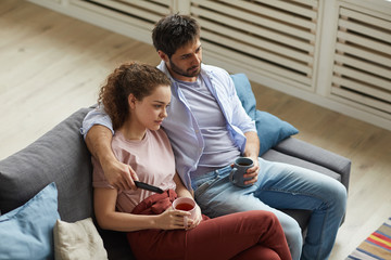 High angle portrait of modern couple watching TV and holding mugs while sitting on sofa at home in cozy apartment and enjoying lazy time, copy space