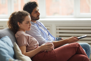 Obraz premium Side view portrait of modern couple watching TV and holding mugs while sitting on sofa at home in cozy apartment, copy space