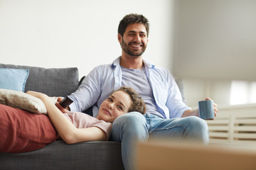Obraz premium Portrait of happy couple watching TV together while sitting on sofa at home in cozy apartment enjoying lazy time, copy space
