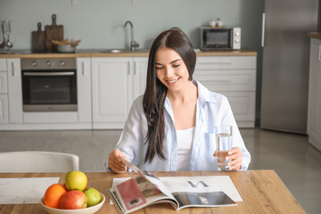 Beautiful young woman drinking water while reading magazine in kitchen
