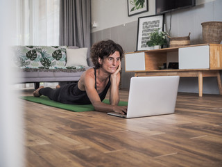 happy and smiling women lying on the floor on her stomach in her living room at home ooking at laptop holding her chin with her hand and typing