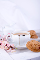 White porcelain cup of tea, milk jug, and fresh-baked oat cookies. English breakfast still-life with drink and treats and tablecloth.