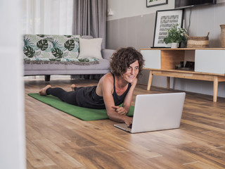women lying on her stomach on a green mat in her living room at home looking at laptop screen getting ready to do an online sport / yoga session