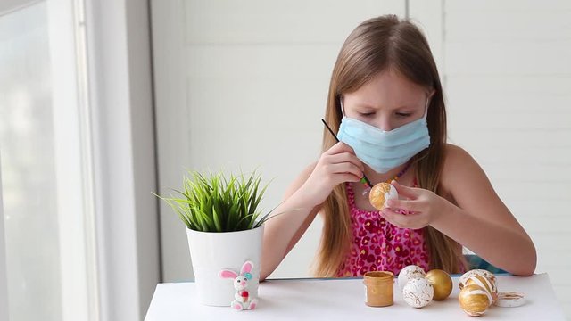 Little girl in a medical mask paints Easter eggs with a pattern. Stay home.