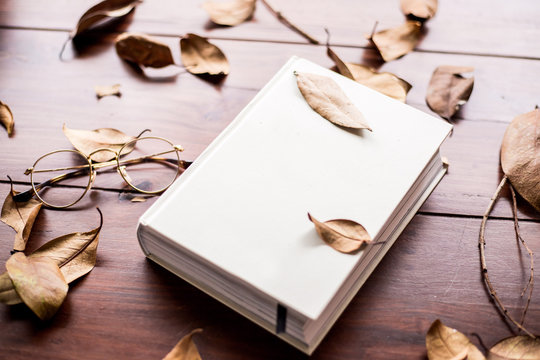 white book on a wooden table with glasses and some leafs