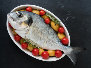 sea bream, tomatos, potatos and rosemary on a serving dish on slate background. ready to cook