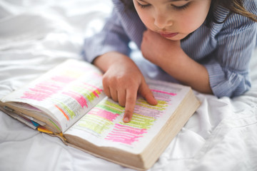 Little girl reading from Bible while lying in bed. Bible with marks on letters.