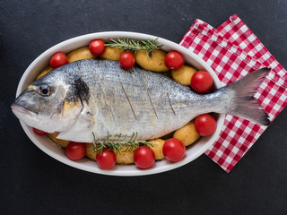 sea bream, tomatos, potatos and rosemary on a serving dish on slate background. ready to cook