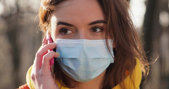 Portrait Woman dressed in orange red coat wearing Protective Face Mask talking mobile phone in the park . close up view. Sunny day Sun flayers pandemic disease virus. COVID-19 coronavirus infection