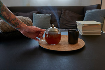 
Steaming teapot with a glass on a black leather table. It is in the living room of a house, with an armchair with cushions behind.