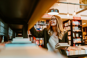 Photo of young blonde woman with glasses looking at book shelves at bookstore. Young woman is...