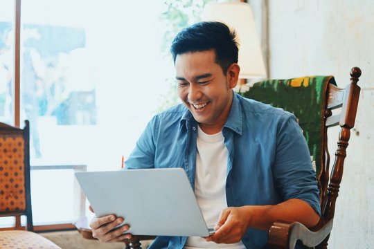 Asian Man In Casual Blue Shirt Working With Laptop From Home.