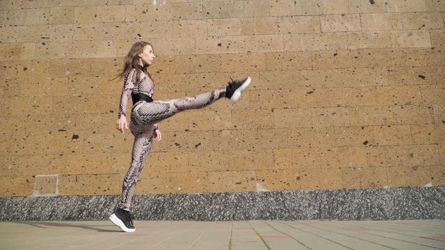 Young Beautiful Athletic Girl Gymnast with Steep Stretch. She goes and does leg in split. Dressed in her slinky jumpsuit with picture scales Cobra. On a brown background concrete wall. Slow Motion