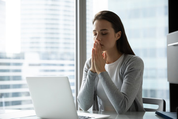 Religious young woman sit at desk with eyes closed hold hands in prayer ask beg about good luck or...