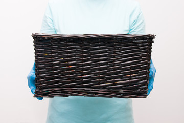 woman in a light blue t-shirt in blue disposable gloves holds a dark brown rectangular basket, contactless delivery concept, light background, quarantine 2020