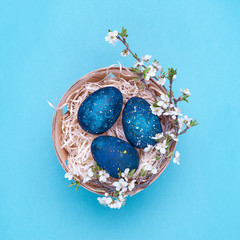 Azure easter eggs in a basket with flowers on a violet background. Square photograph. Top view