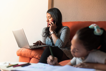 Latin young woman working at home calling with the mobile phone using the laptop while her daughter...