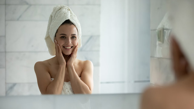 Smiling young woman in bath towel look in mirror after shower satisfied with healthy glowing facial skin, happy millennial girl touch clean face after beauty procedures or treatment, skincare concept