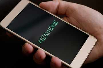 Hashtag stay home! The inscription on the smartphone screen. Male hand holds a phone on a dark background. The recommendation is quarantined at home to prevent coronovirus covid 19.