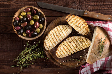 Sliced traditional toast bread with olives, garlic and herbs on a cutting board and dark table