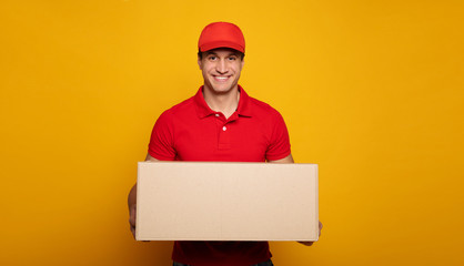 Handsome young happy courier or delivery man in red uniform is posing isolated on yellow background.