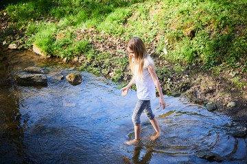 Child cute blond girl playing in the creek. Girl walking barefoot in forest stream  and exploring nature. Summer children fun. Children summer activities
