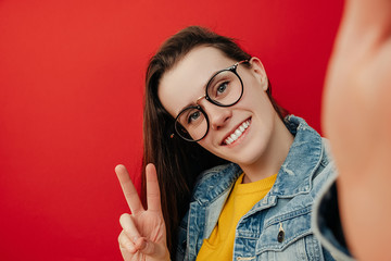 Cheerful young woman in eyeglasses, makes peace gesture, takes selfie on smartphone, has fun with cellular, wears denim jacket, poses against red background with blank copy