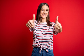 Young beautiful brunette woman wearing casual striped t-shirt standing over red background approving doing positive gesture with hand, thumbs up smiling and happy for success. Winner gesture.