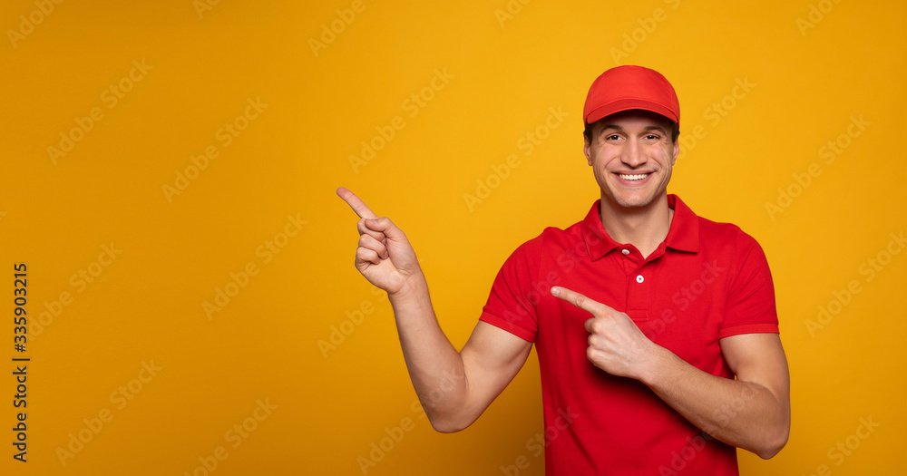 Wall mural Handsome young happy courier or delivery man in red uniform is posing isolated on yellow background.