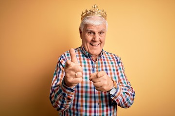 Senior handsome hoary man wearing golden crown of king over isolated yellow background pointing fingers to camera with happy and funny face. Good energy and vibes.
